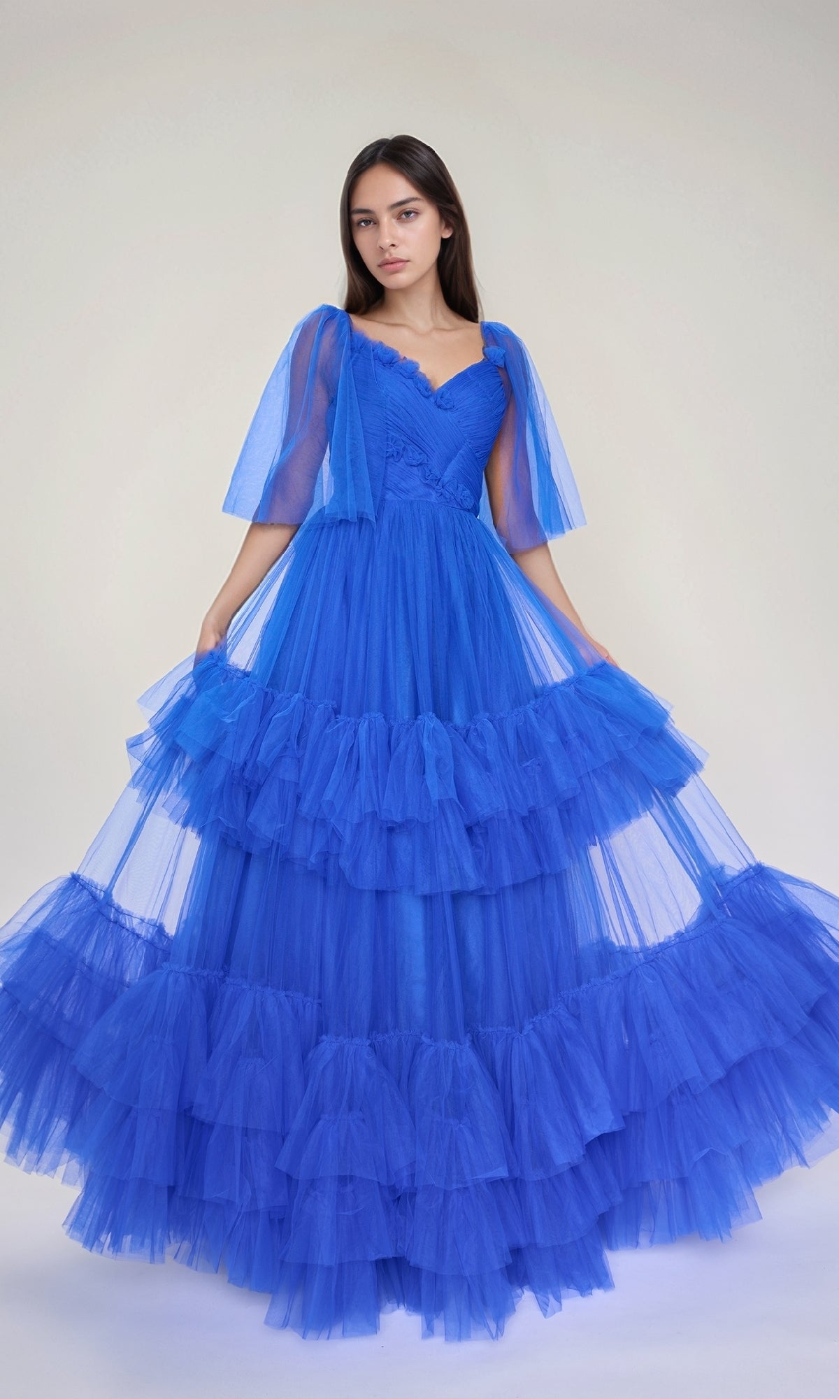 A woman models the Nina Canacci 5315, a bright blue, floor-length long formal dress with ruffle tiered tulle and sheer short sleeves, posing against a plain background.